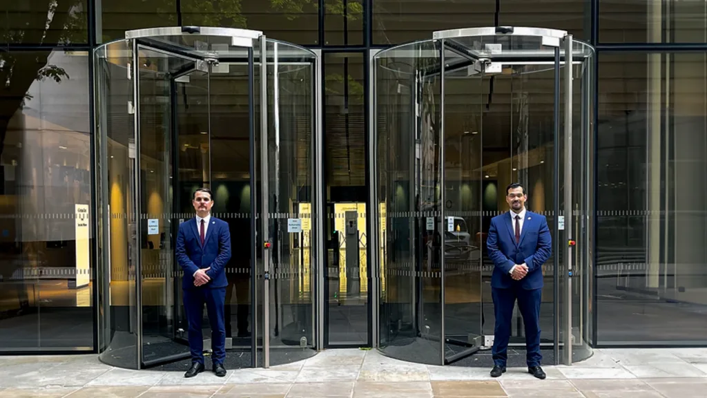 Two hotel security guards, dressed in Guard Mark uniforms, stand by the glass door, diligently performing their duties.