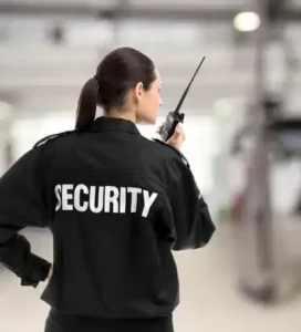 Female security guard in black uniform with "SECURITY" on back, using walkie-talkie in a spacious indoor area.