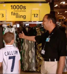 Retail security guard assisting a young boy named Callum in a retail clothing section with a sale sign overhead.