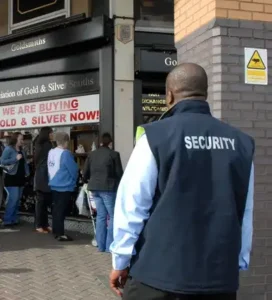 Male Retail security guard in a blue uniform with "SECURITY" on back, monitoring a crowd outside a jewelry store.