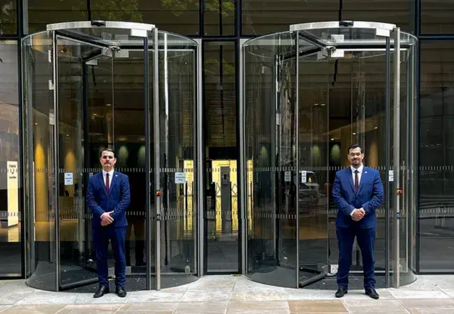 Two hotel security guards, dressed in Guard Mark uniforms, stand by the glass door, diligently performing their duties.