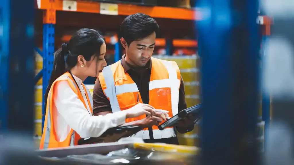 Two warehouse workers wearing orange safety vests review information on a tablet in a storage facility.