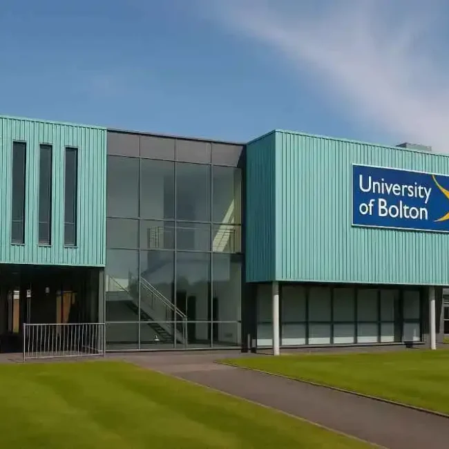 Front view of a modern University of Bolton campus building with large glass windows, teal cladding, and the university’s logo on display, surrounded by green lawns under a clear blue sky.