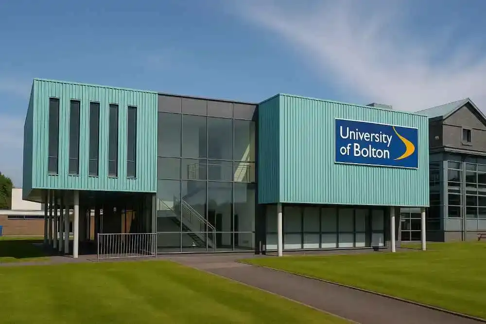 Front view of a modern University of Bolton campus building with large glass windows, teal cladding, and the university’s logo on display, surrounded by green lawns under a clear blue sky.